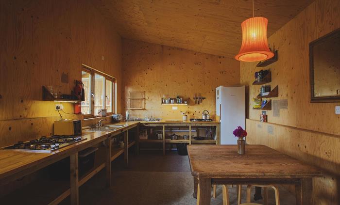 An indoor camp kitchen with a large kitchen space with countertops and hobs. There is a table in the foreground with a light above. A mirror hangs on